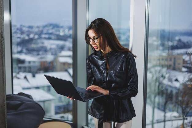 Mulher jovem freelancer em óculos com laptop trabalhando remotamente no espaço de trabalho moderno com grandes janelas Trabalho remoto