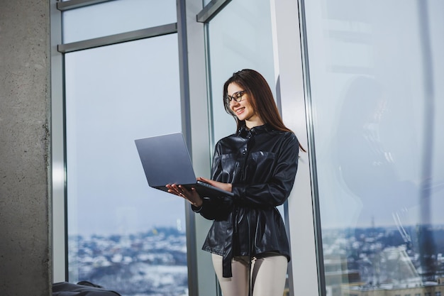 Mulher jovem freelancer em óculos com laptop trabalhando remotamente no espaço de trabalho moderno com grandes janelas Trabalho remoto