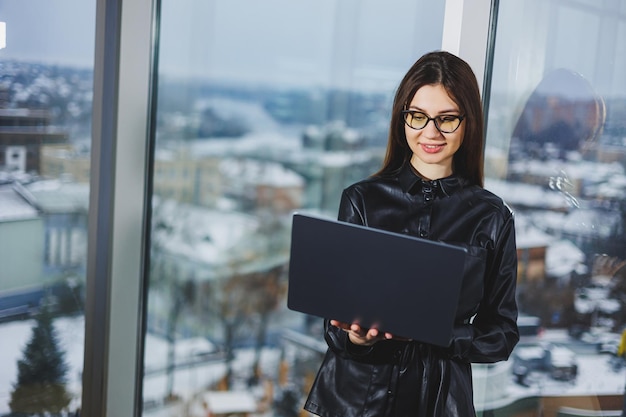 Mulher jovem freelancer em óculos com laptop trabalhando remotamente no espaço de trabalho moderno com grandes janelas Trabalho remoto