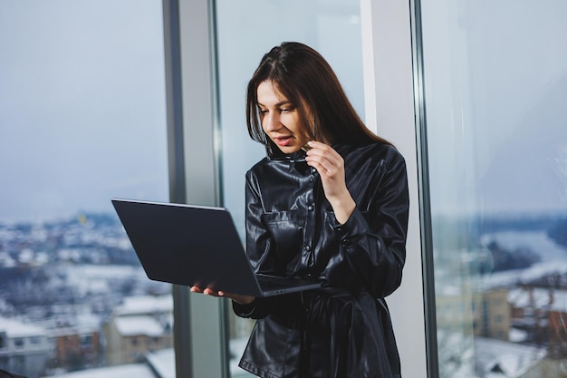 Mulher jovem freelancer em óculos com laptop trabalhando remotamente no espaço de trabalho moderno com grandes janelas Trabalho remoto