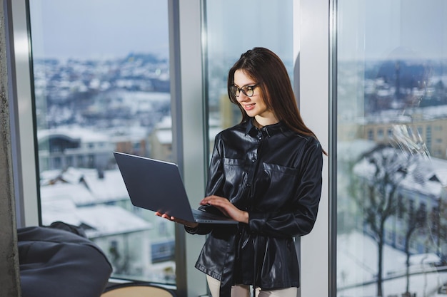 Mulher jovem freelancer em óculos com laptop trabalhando remotamente no espaço de trabalho moderno com grandes janelas Trabalho remoto