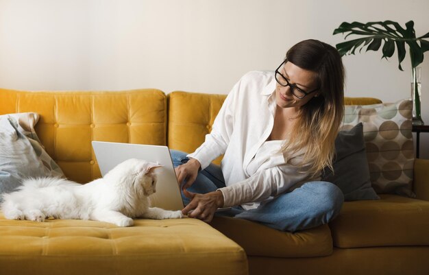 Mulher jovem freelancer usando óculos sentado no sofá amarelo dentro de casa trabalhando com laptop e celular