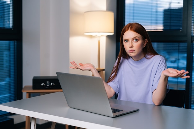 Mulher jovem frustrada, confusa com o problema do computador laptop, sentada na mesa do escritório em casa perto da janela à noite, tarde da noite, olhando para a câmera.