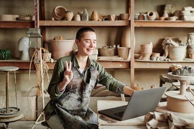 Mulher jovem gesticulando e sorrindo enquanto fala online no laptop durante seu trabalho em um estúdio de cerâmica