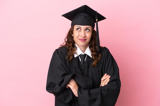 Mulher jovem graduada da universidade isolada em fundo rosa fazendo gesto de dúvidas enquanto levanta os ombros