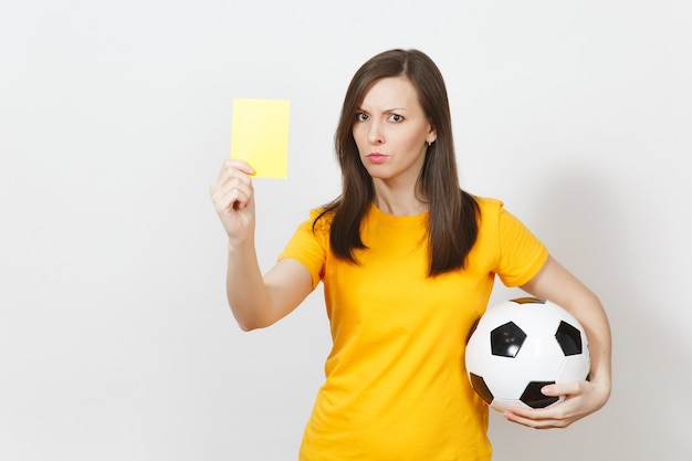 Mulher jovem grave grave europeia, árbitro de futebol ou jogador de uniforme amarelo, mostrando o cartão amarelo, segurando uma bola de futebol isolada no fundo branco. Esporte joga futebol, conceito de estilo de vida saudável.