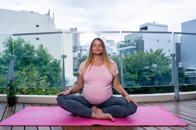 Mulher jovem grávida em casa praticando ioga e meditação em um tapete rosa