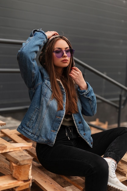 Mulher jovem hippie na moda em óculos roxos da moda em jaqueta jeans elegante azul em jeans vintage preto posando em paletes de madeira na cidade. Menina muito elegante do modelo de moda relaxa ao ar livre.