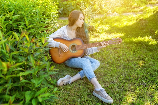 Mulher jovem hippie sentada na grama tocando guitarra no parque ou jardim