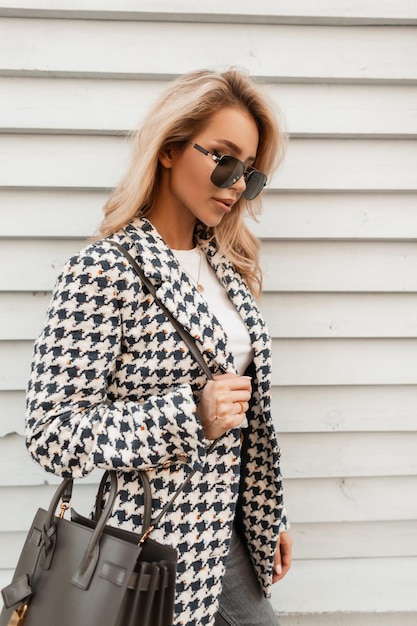 Mulher jovem hipster loira na moda com óculos de sol em roupas de moda com jaqueta e bolsa fica perto da parede de madeira