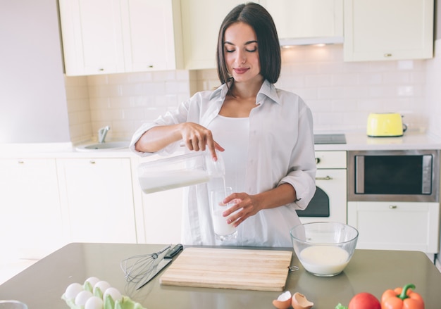 Mulher jovem incrível fica na cozinha e derramando leite no copo.