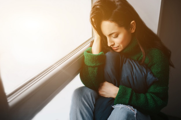 Mulher jovem infeliz é triste perto da janela. A morena está chorando. Tristeza, apatia, depressão e melancolia.