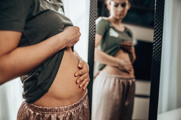 Mulher jovem infeliz em frente a um espelho e segurando as mãos em sua barriga inchada.