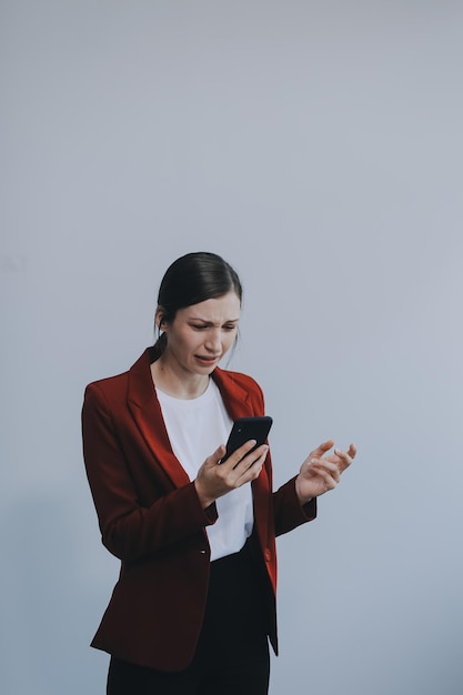 Mulher jovem irritada irritada com mensagem de spam, telefone preso, olhando para o smartphone, isolada em fundo de estúdio em branco, adolescente furiosa com problema com o telemóvel, irritada pelo telemóvil quebrado.