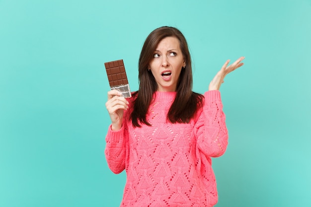 Mulher jovem irritada no suéter rosa de malha, olhando para cima, espalhando as mãos, segurar a barra de chocolate isolada no fundo da parede azul turquesa, retrato de estúdio. Conceito de estilo de vida de pessoas. Simule o espaço da cópia.