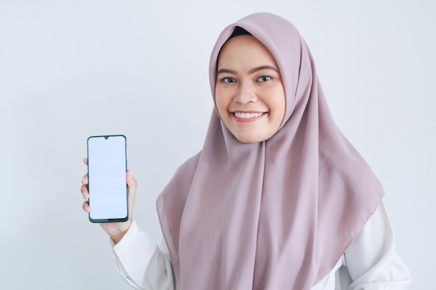 Mulher jovem islâmica asiática usando lenço na cabeça está apontando o dedo na tela branca do telefone com sorriso e sentimento feliz mulher indonésia em fundo cinza