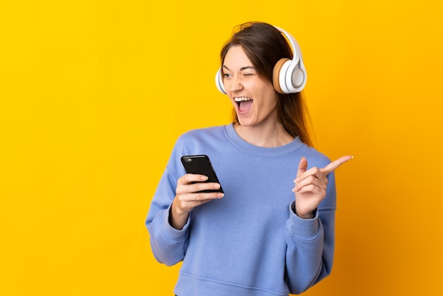 Mulher jovem isolada na parede amarela ouvindo música com um celular e cantando