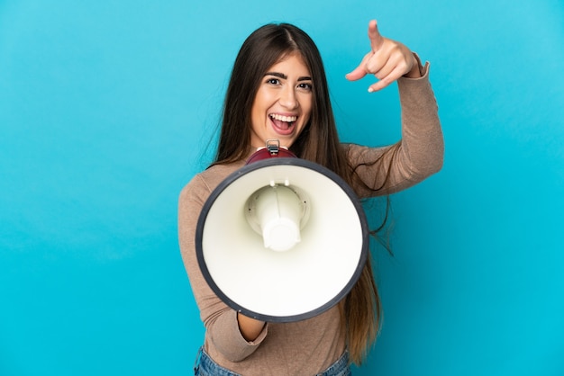 Mulher jovem isolada na parede azul gritando em um megafone para anunciar algo enquanto aponta para a frente