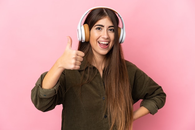 Mulher jovem isolada na parede rosa ouvindo música e com o polegar para cima