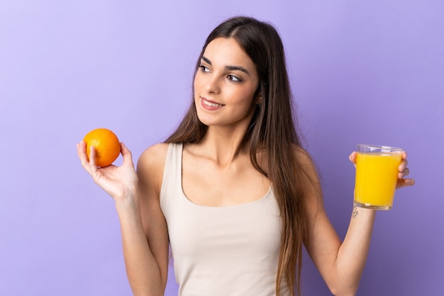 Mulher jovem isolada na parede roxa segurando uma laranja e um suco de laranja