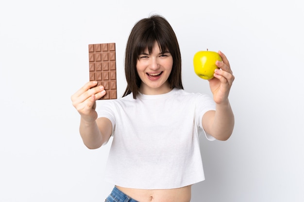 Mulher jovem isolada no branco pegando um comprimido de chocolate com uma das mãos e uma maçã com a outra