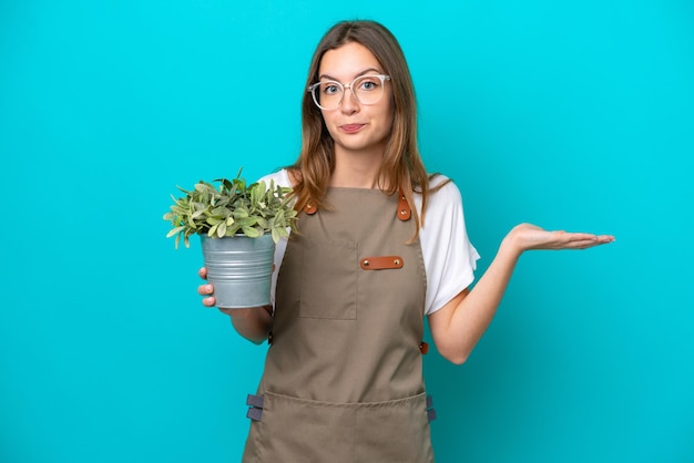 Mulher jovem jardineiro caucasiano segurando uma planta isolada em fundo azul com dúvidas ao levantar as mãos