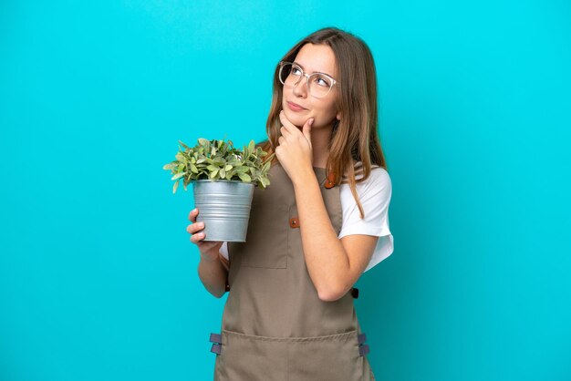 Mulher jovem jardineiro caucasiano segurando uma planta isolada em fundo azul com dúvidas