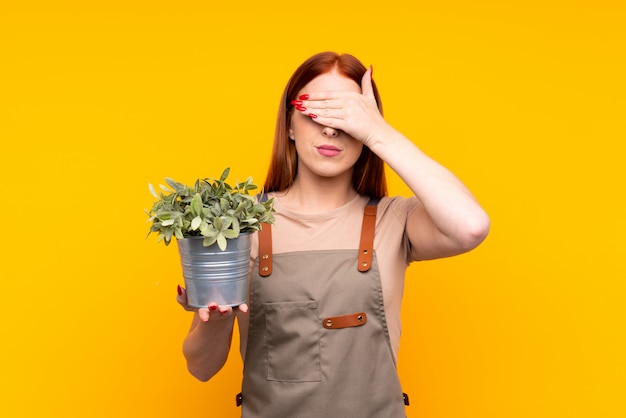 Mulher jovem jardineiro ruiva segurando uma planta cobrindo os olhos pelas mãos