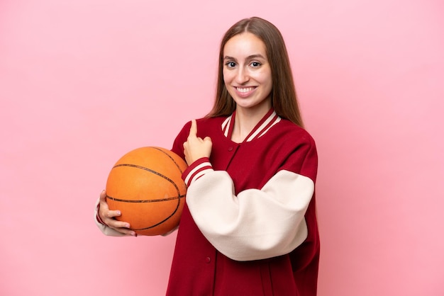 Mulher jovem jogador de basquete caucasiano isolada sobre fundo rosa apontando para trás