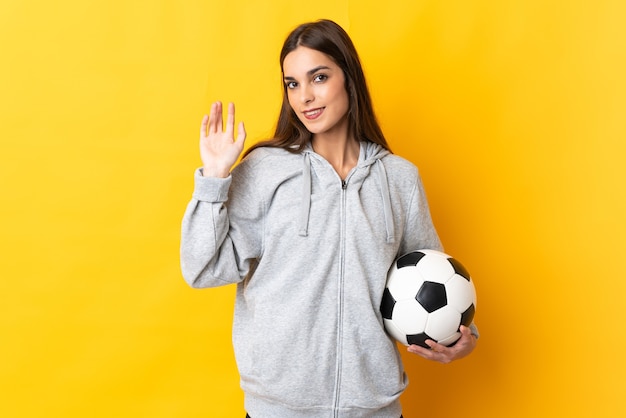 Mulher jovem jogador de futebol isolada em fundo amarelo saudando com a mão com expressão feliz