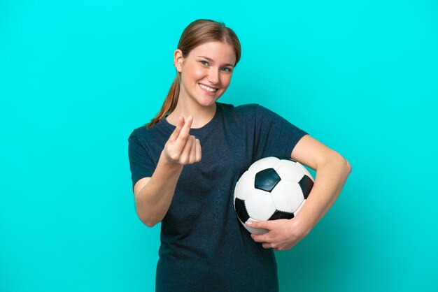 Mulher jovem jogador de futebol isolada em fundo azul, fazendo gesto de dinheiro