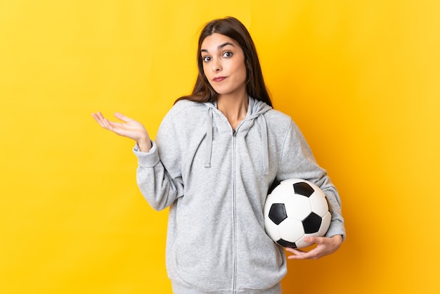 Mulher jovem jogador de futebol isolada na parede amarela tendo dúvidas ao levantar as mãos