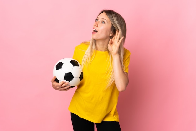 Mulher jovem jogador de futebol isolada na parede rosa ouvindo algo