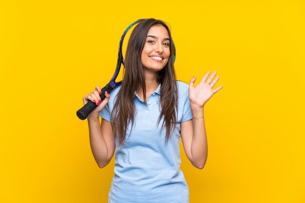 Mulher jovem jogador de tênis sobre parede amarela isolada, saudando com mão com expressão feliz