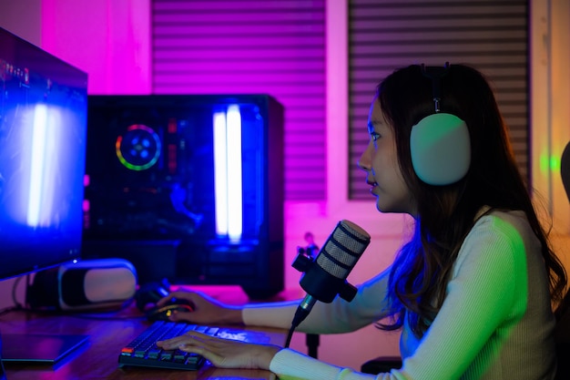 Mulher jovem jogadora asiática usa fones de ouvido jogando videogame on-line no computador, ela transmite ao vivo e conversa com os fãs, Fêmea feliz falando com microfone na sala de jogos, conceito de streaming de jogos