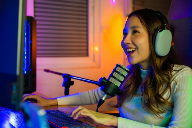 Mulher jovem jogadora asiática usa fones de ouvido jogando videogame on-line no computador, ela transmite ao vivo e conversa com os fãs, fêmea feliz falando com microfone na sala de jogos, conceito de streaming de jogos