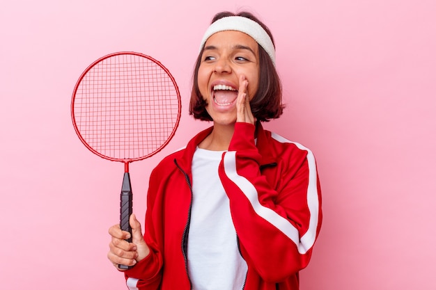 Mulher jovem jogando badminton isolada na parede rosa, gritando e segurando a palma da mão perto da boca aberta