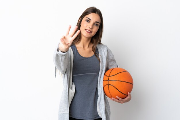 Mulher jovem jogando basquete isolada na parede branca feliz e contando três com os dedos