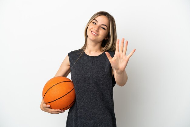 Mulher jovem jogando basquete sobre uma parede branca isolada, contando cinco com os dedos