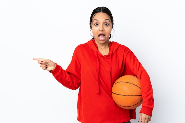 Mulher jovem jogando basquete sobre uma parede branca isolada surpresa e apontando para o lado