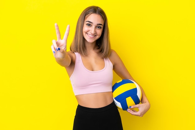 Mulher jovem jogando vôlei isolada em um fundo amarelo sorrindo e mostrando sinal de vitória