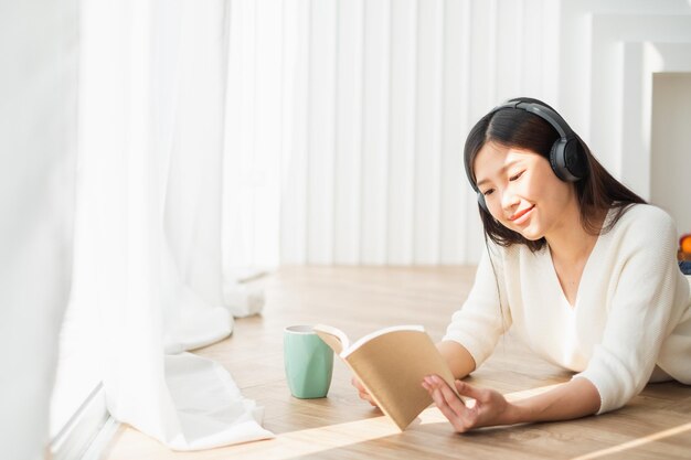 Mulher jovem lendo um livro enquanto ouve música por uma xícara de café no chão