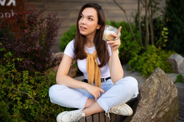 Mulher jovem livre encantadora em look casual, bebendo café na rua.