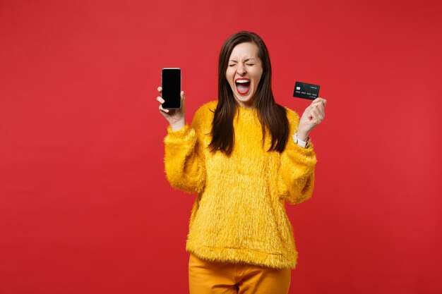 Mulher jovem louca com os olhos fechados, gritando, segurando o telefone móvel com a tela em branco e vazia, cartão de banco de crédito isolado sobre fundo vermelho. Emoções sinceras de pessoas, conceito de estilo de vida. Simule o espaço da cópia.