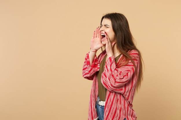 Mulher jovem louca em roupas casuais, de olhos fechados, gritando com as mãos perto da boca isolada em fundo bege pastel em estúdio. Emoções sinceras de pessoas, conceito de estilo de vida. Simule o espaço da cópia.