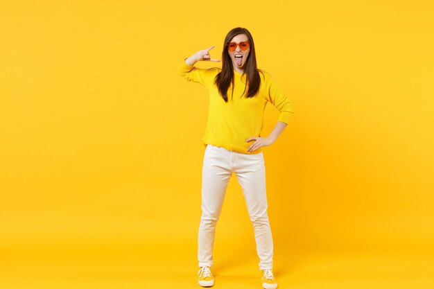 Mulher jovem louca em roupas casuais, mostrando a língua, retratando o sinal de rock heavy metal isolado no fundo da parede laranja amarela no estúdio. Emoções sinceras de pessoas, conceito de estilo de vida. Mock up espaço de cópia.