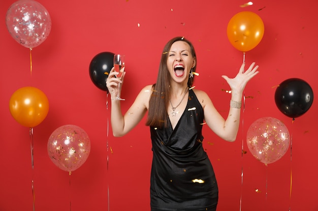 Mulher jovem louca em um vestido preto gritando comemorando, segurando a taça de champanhe e pegando confetes em balões de ar de fundo vermelho brilhante. Feliz Ano Novo, conceito de festa de feriado de maquete de aniversário.