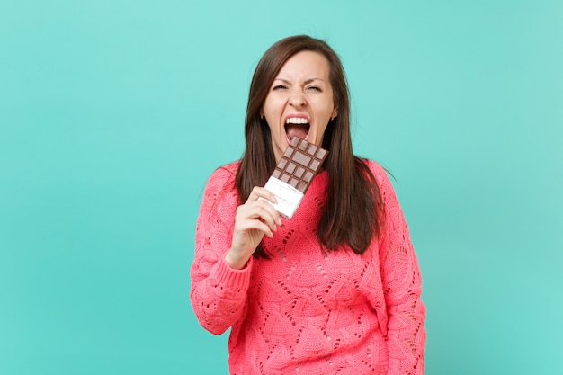 Mulher jovem louca no suéter rosa de malha, segurando na mão, mordendo a barra de chocolate, gritando, isolado no fundo da parede azul turquesa, retrato de estúdio. conceito de estilo de vida de pessoas. simule o espaço da cópia.