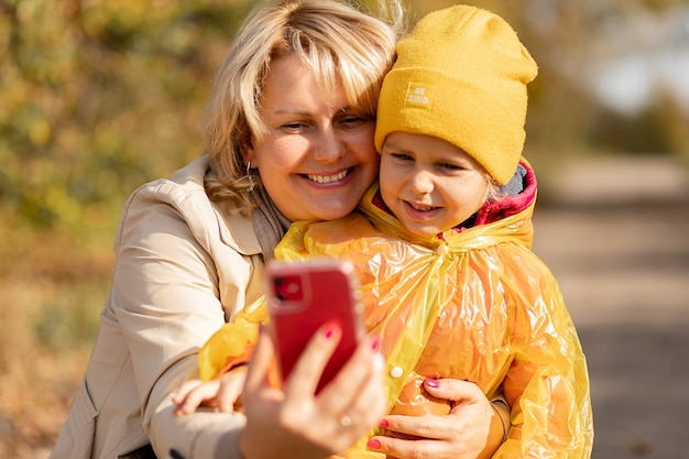 Mulher jovem, mãe de uma menina, usa o smartphone para fazer videochamada para parentes ou tira uma selfie no meio do parque de outono