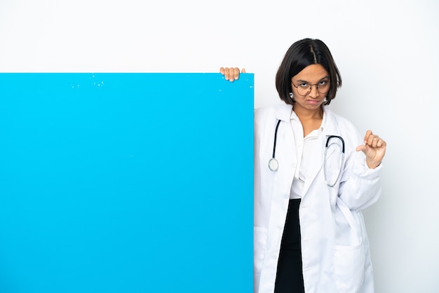 Mulher jovem médica de raça mista com um grande cartaz isolado no fundo branco mostrando o polegar para baixo com expressão negativa